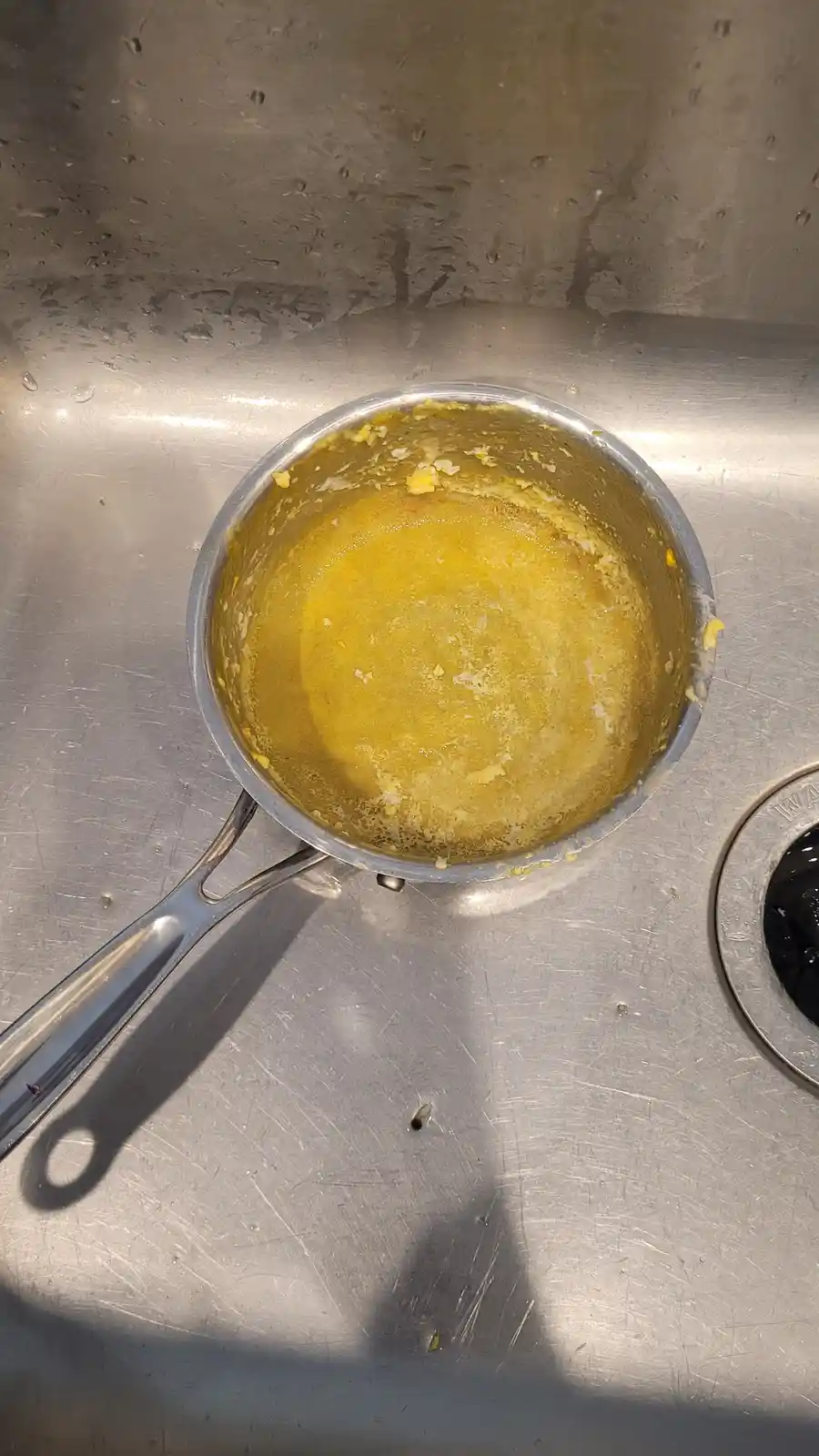 A stainless steel saucepan sits in a stainless steel sink, containing a thick, yellow, lumpy substance with bits of solid…