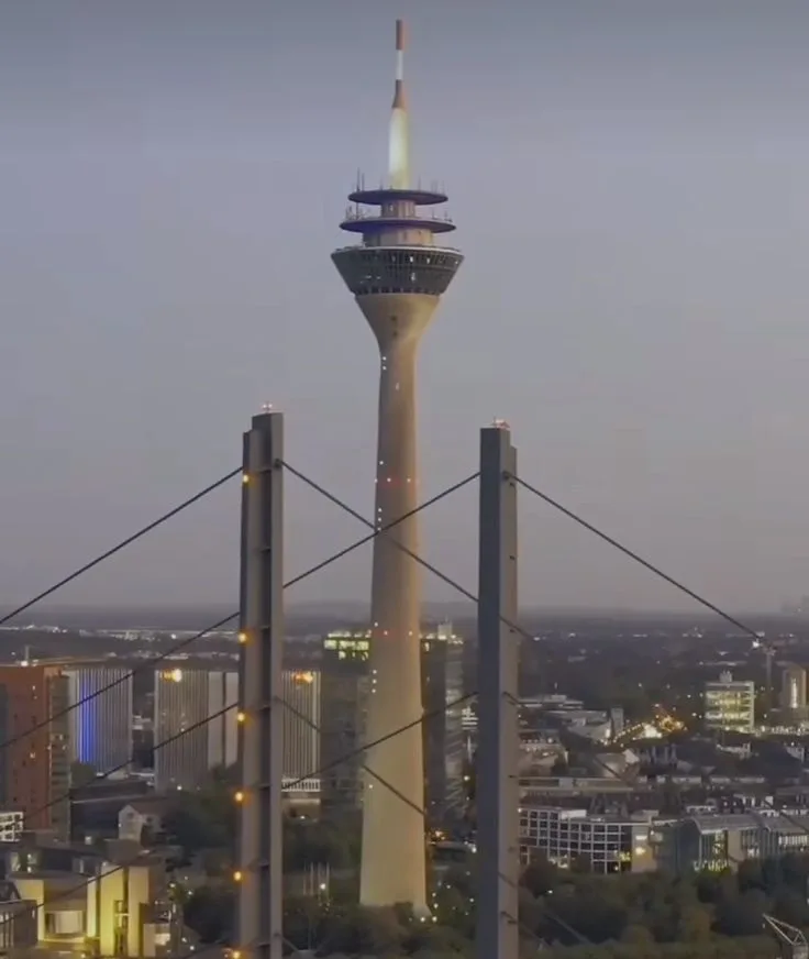A tall, slender tower with a lit observation deck and antenna stands in a cityscape at dusk, framed by the cables and pylons…