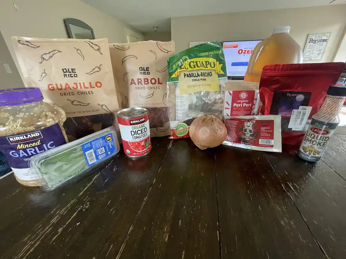 A dark wooden table displays an array of spicy cooking ingredients including bags of Ole Rico Guajillo and Arbol dried…
