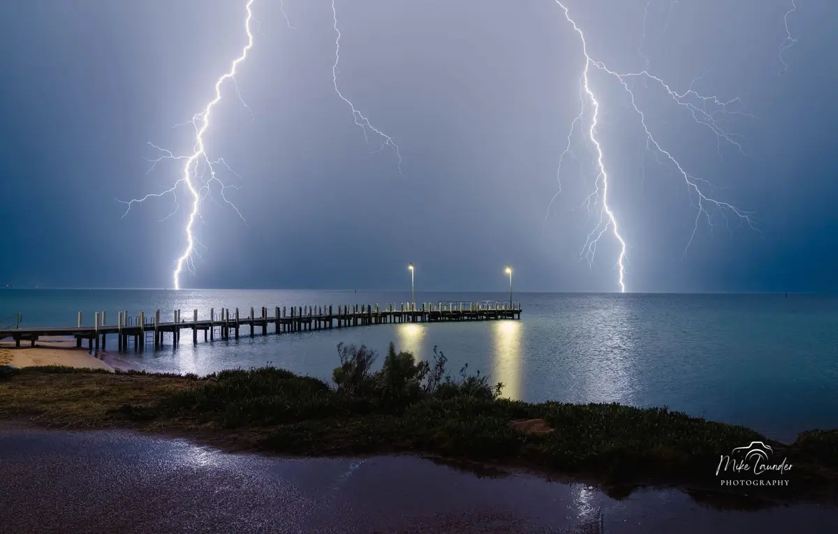 A dramatic photograph shows two bright lightning bolts striking the ocean at night, illuminating a wooden pier with two lit…