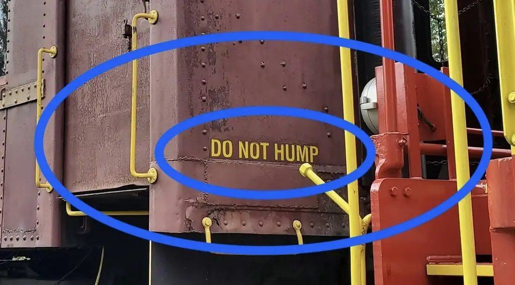 A close-up photo of a rusty, riveted metal train car side with yellow safety rails, featuring the text 'DO NOT HUMP' in…