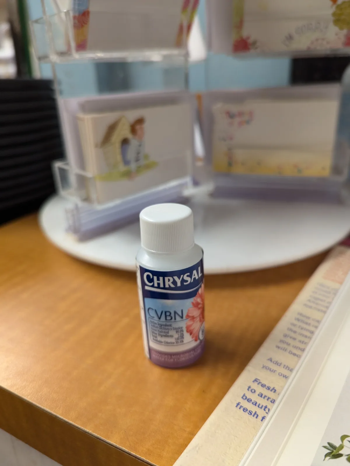 A small bottle labeled 'CHRY SAL CVBN' sits on a wooden counter, with floral greeting cards in a rack behind it. The…