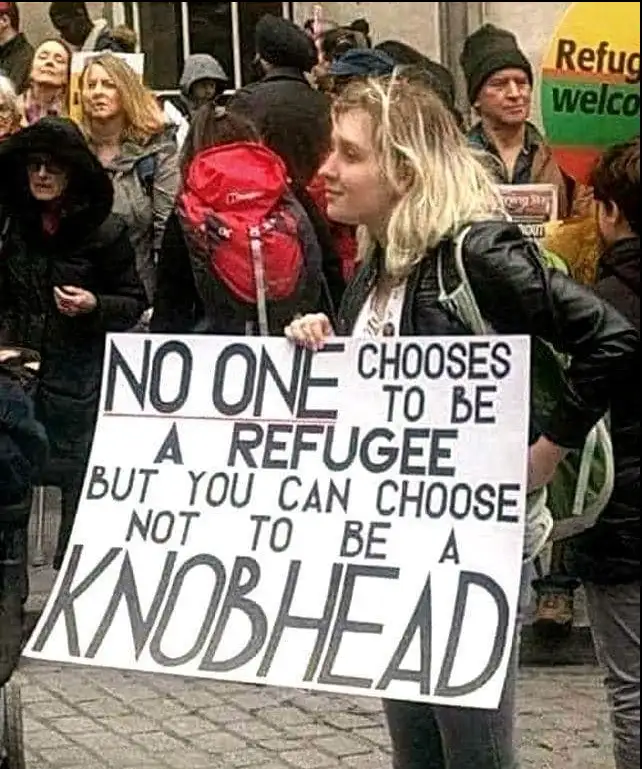 A woman at a protest holds a large white sign with bold black text stating 'NO ONE CHOOSES TO BE A REFUGEE BUT YOU CAN…