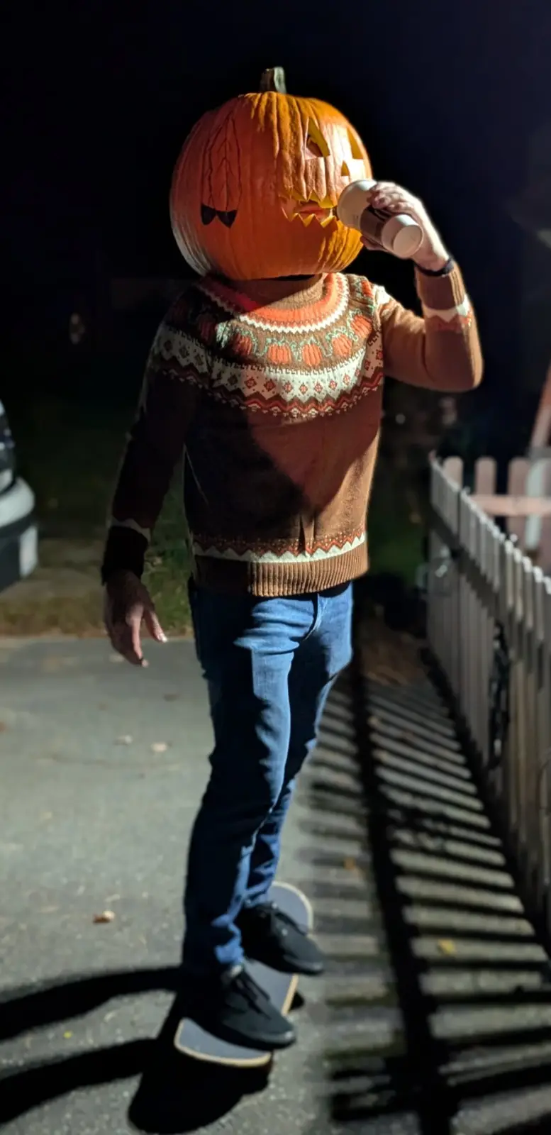 A person wearing a carved jack-o'-lantern for a head, a festive sweater, and jeans, is standing on a skateboard at night…