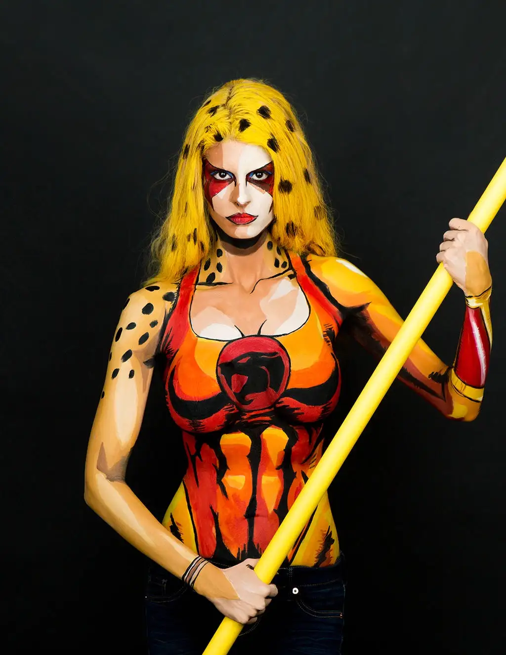 A woman with yellow hair and black spots is painted to resemble the character Cheetah from DC Comics, holding a yellow staff…