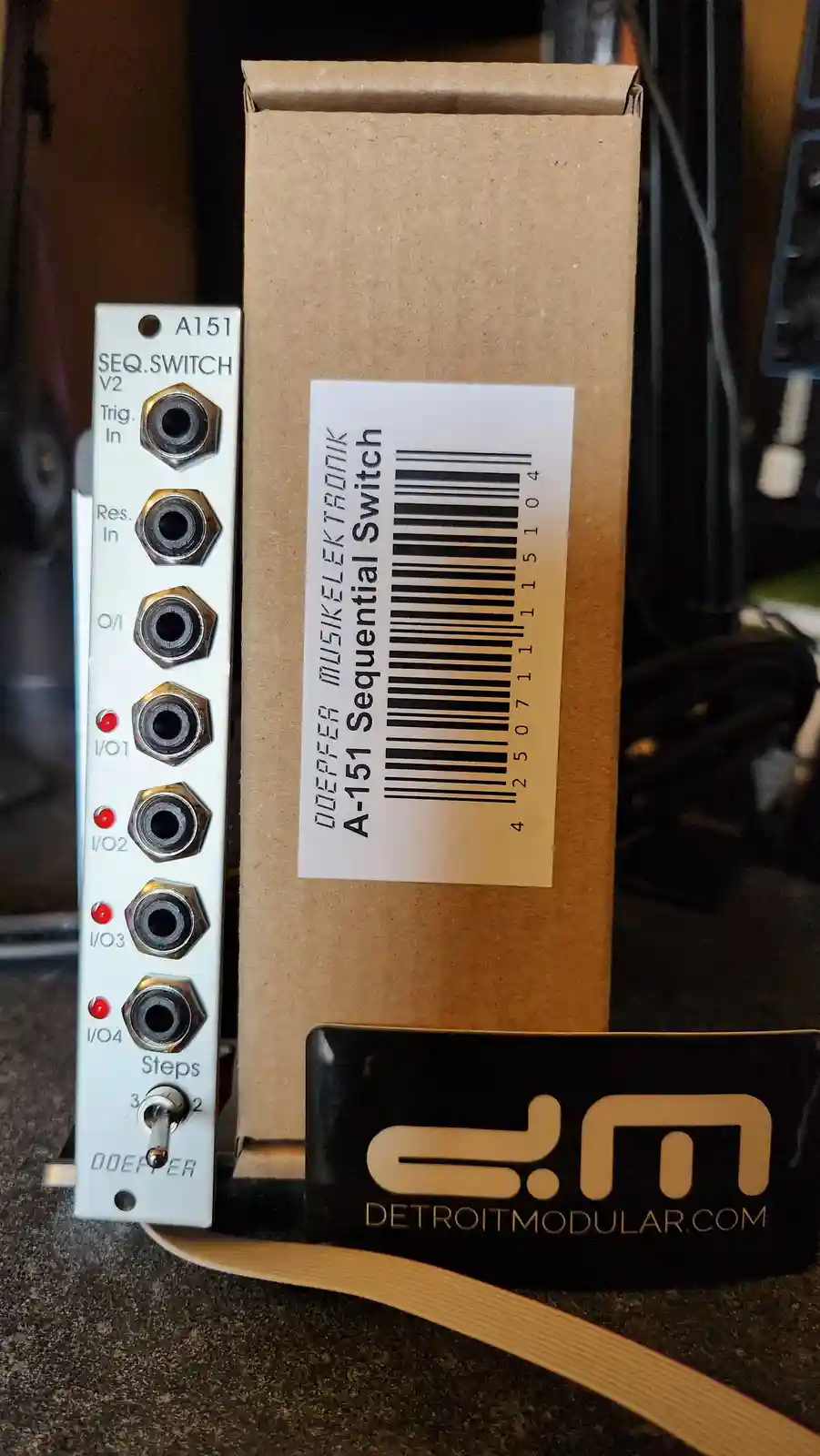 A white modular synthesizer module labeled 'A151 SEQ.SWITCH V2' with multiple jacks and red LEDs sits next to its brown…