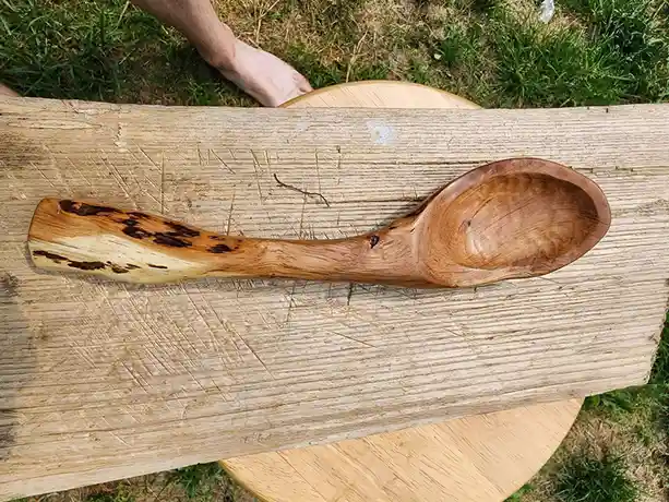 A hand-carved wooden spoon with a rustic, natural finish rests on a weathered wooden plank outdoors. The spoon's handle…
