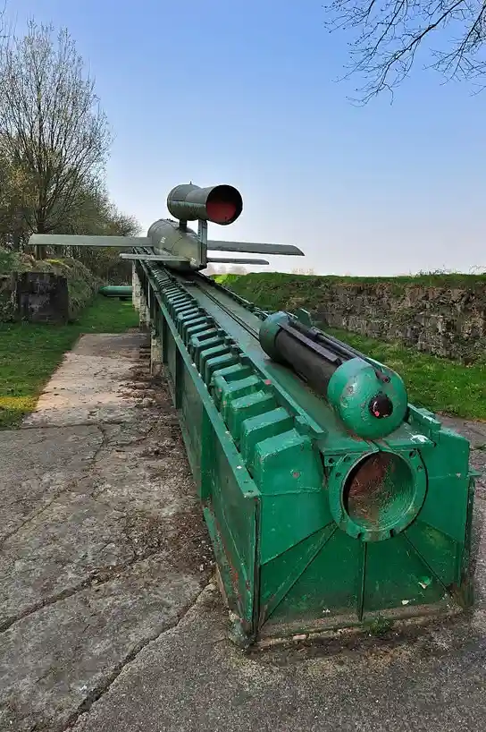 A green, weathered military missile launcher with a missile mounted on top sits outdoors on a concrete path, surrounded by…