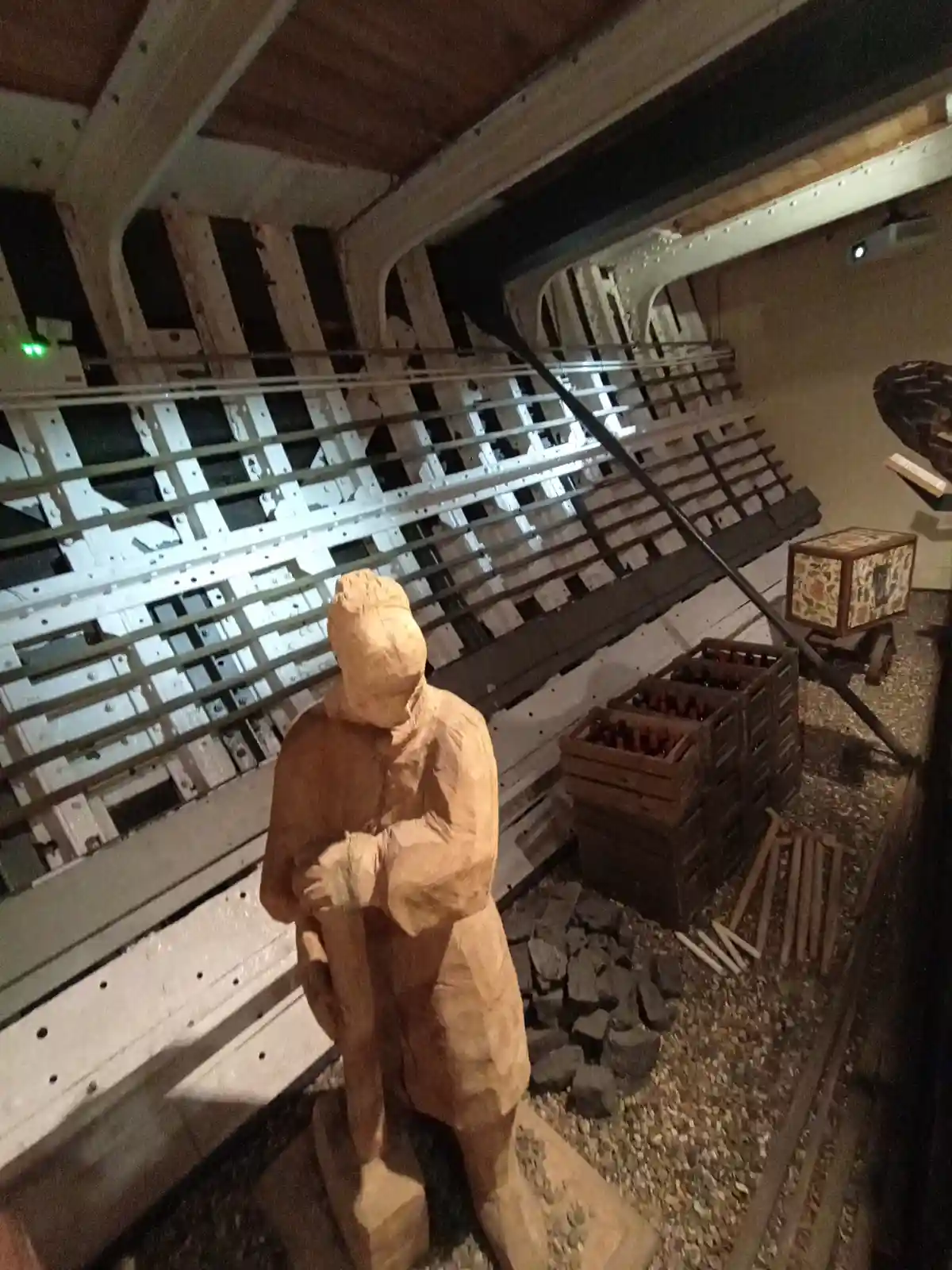 A wooden sculpture of a person in a hooded coat stands inside a model of a ship's hold, surrounded by crates of bottles…