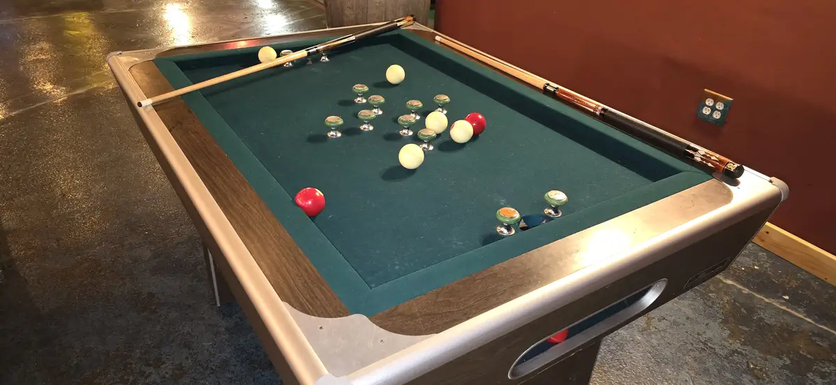 A pool table with green felt and wooden rails has its balls replaced with decorative cabinet knobs, and pool cues rest on…
