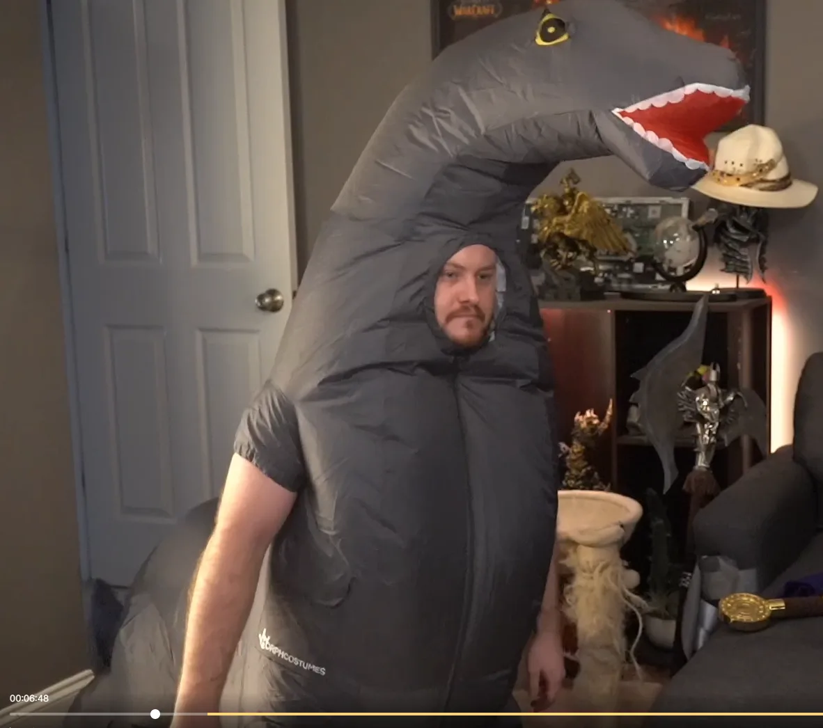A man wearing a large, grey inflatable dinosaur costume with a long neck and open mouth stands in a room. The costume covers…
