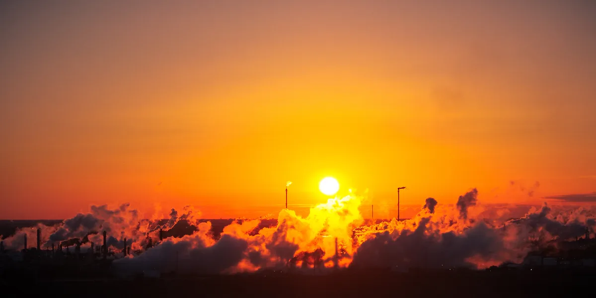 A wide-angle photograph captures a dramatic sunset where the bright orange sun is positioned behind a sprawling industrial…