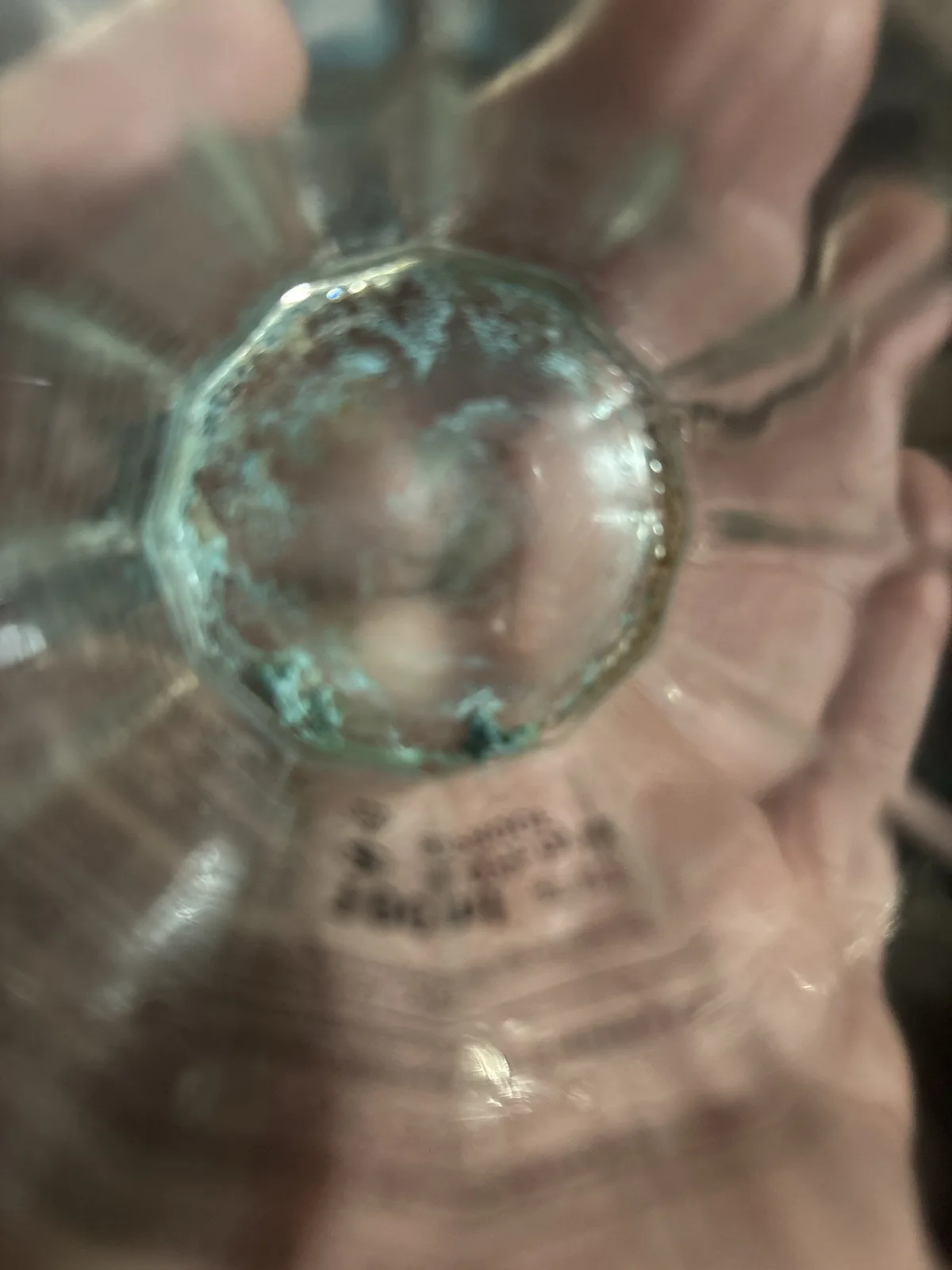 A blurry, close-up photo shows a hand holding a clear glass bottle, viewed from the bottom up. The bottle's base has a…