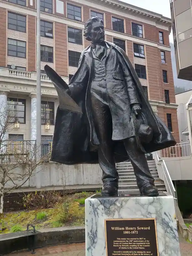 A bronze statue of William Henry Seward stands in front of the Alaska State Capitol, holding a document and a hat. The…