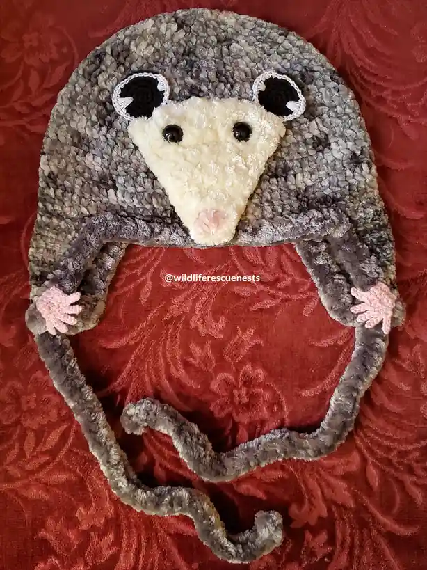 A handmade, crocheted hat shaped like an opossum, featuring a fuzzy gray body, a cream-colored face with black eyes and a…