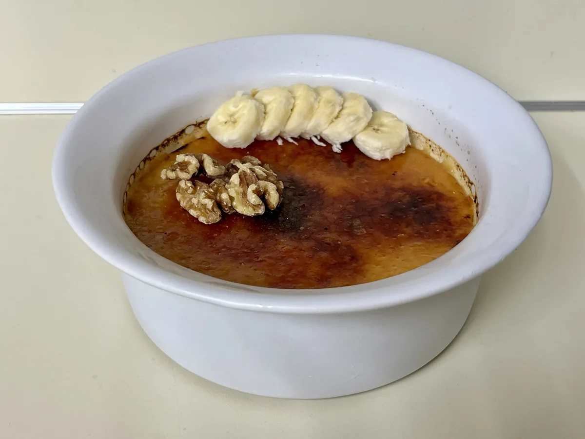 A white ceramic ramekin holds a golden-brown crème brûlée with a caramelized sugar crust. On top are a row of sliced bananas…