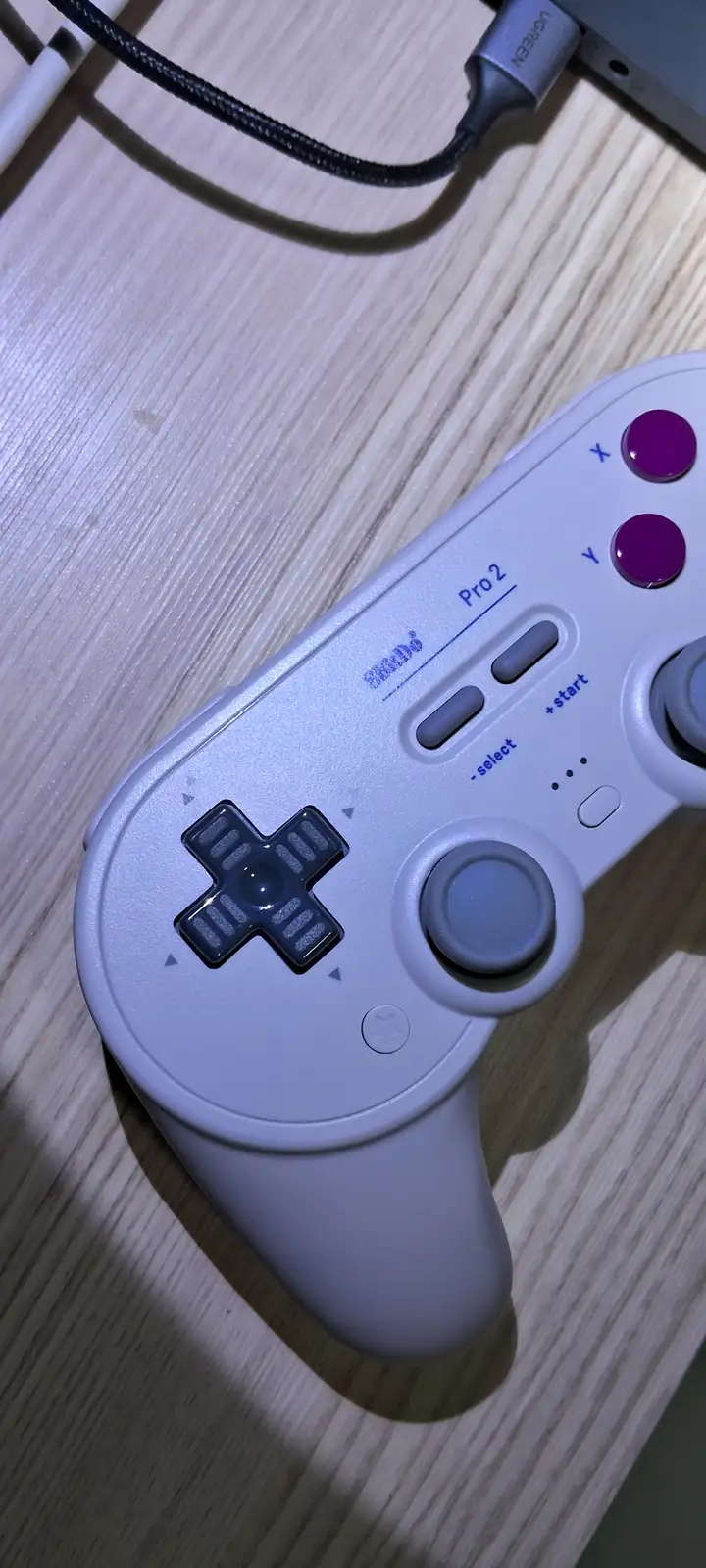 A close-up photo of a white Nintendo Switch Pro 2 controller resting on a wooden surface, showing its directional pad…