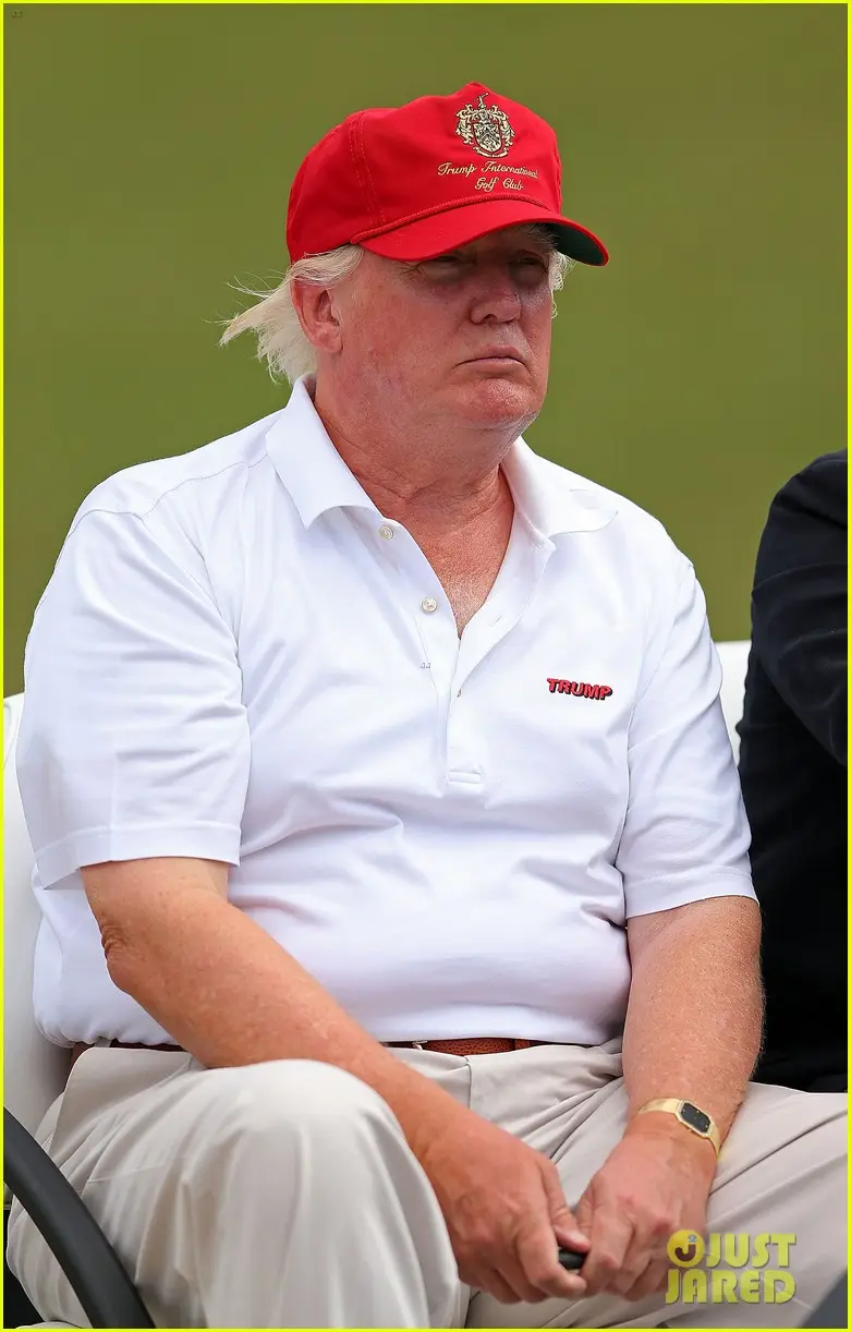 A man with white hair wearing a red 'Trump International Golf Club' cap and a white polo shirt with 'TRUMP' embroidered in…