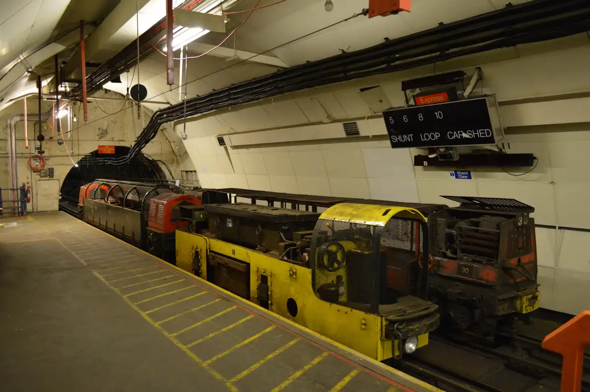 An underground railway maintenance depot shows a yellow and red shunting train stopped at a platform with yellow safety…