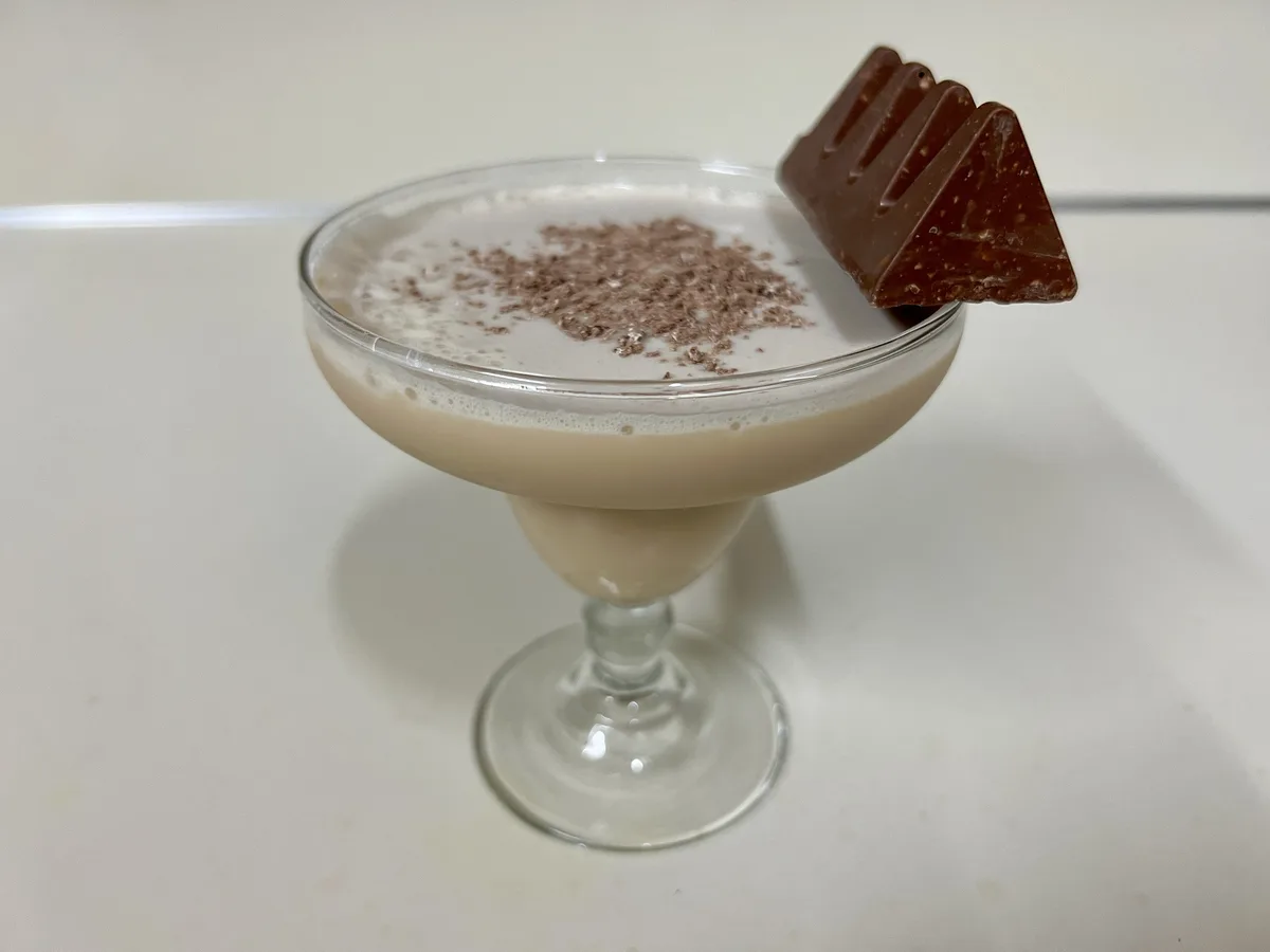 A creamy beige beverage in a stemmed cocktail glass is topped with chocolate shavings and a large, triangular piece of…