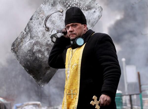 A man dressed as a priest in a black coat and yellow vest with a cross necklace and a gold cross in his hand, holding a…