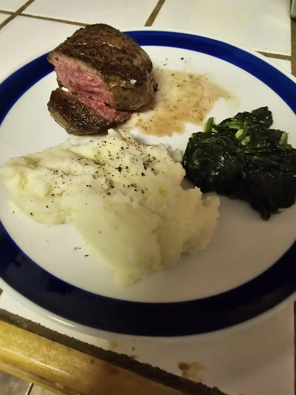 A plate of food featuring a medium-rare steak, mashed potatoes with black pepper, and sautéed spinach, all served on a white…