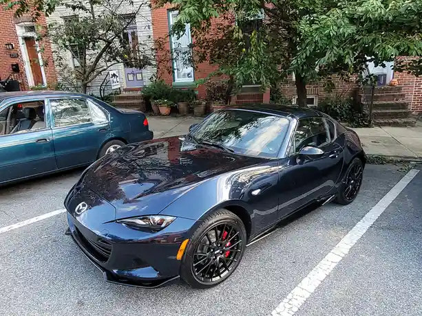 A glossy black Mazda Miata sports car is parked in a residential street, next to a blue sedan. The Miata has black wheels…