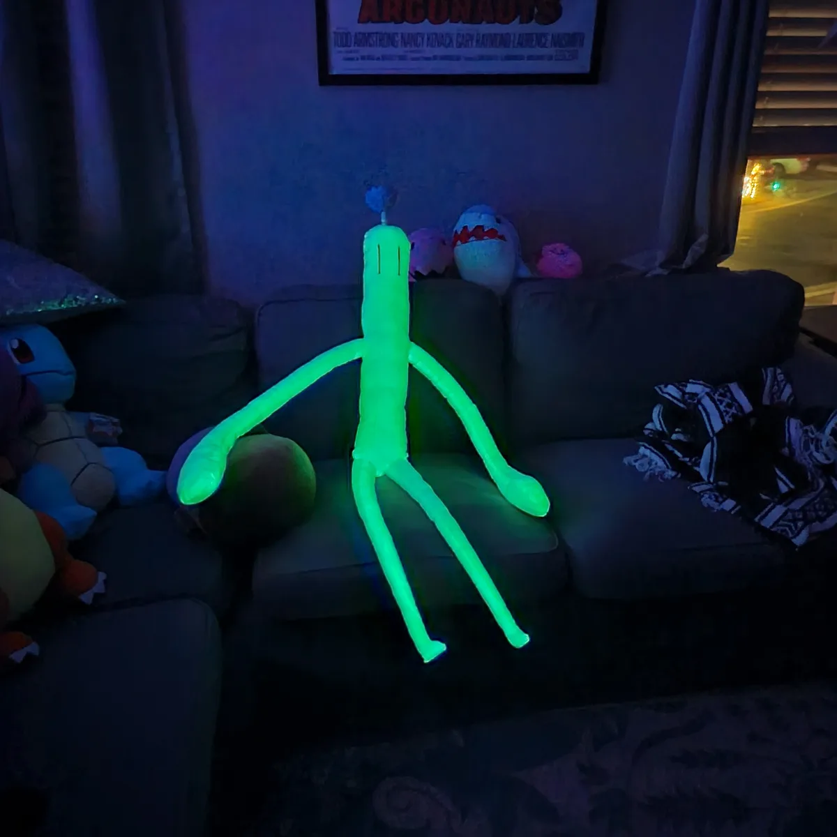 A large, glowing green plush toy resembling the character 'Squidward' from SpongeBob SquarePants sits on a dark couch in a…