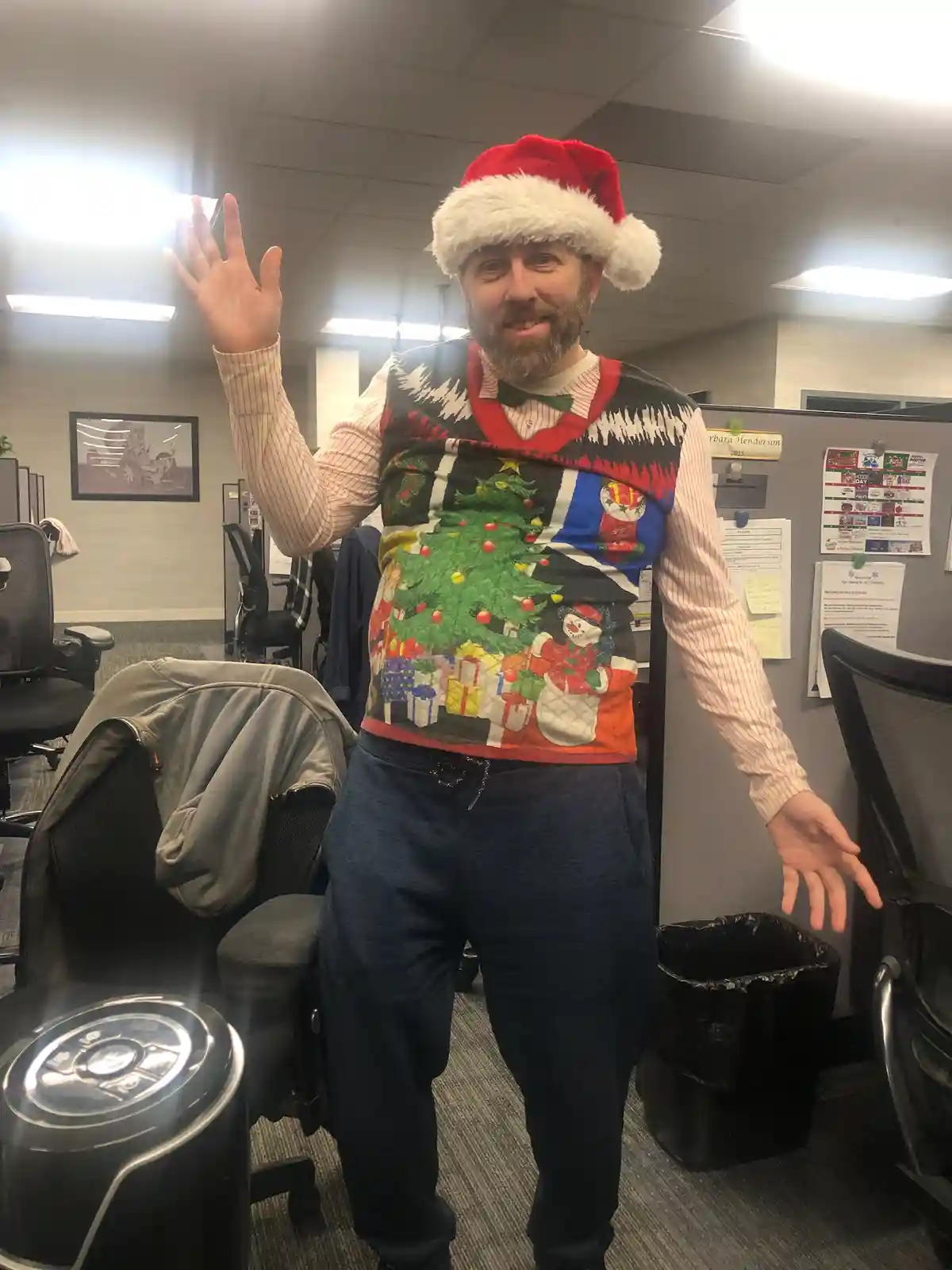 A man with a beard wearing a red and white Santa hat and a very colorful, busy Christmas-themed sweater stands in an office…
