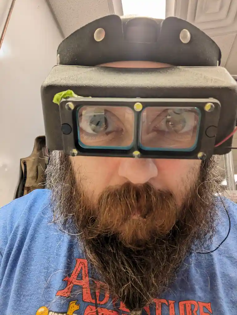 A man with a long beard and mustache wears a large, bulky, custom-built head-mounted device with two lenses covering his…