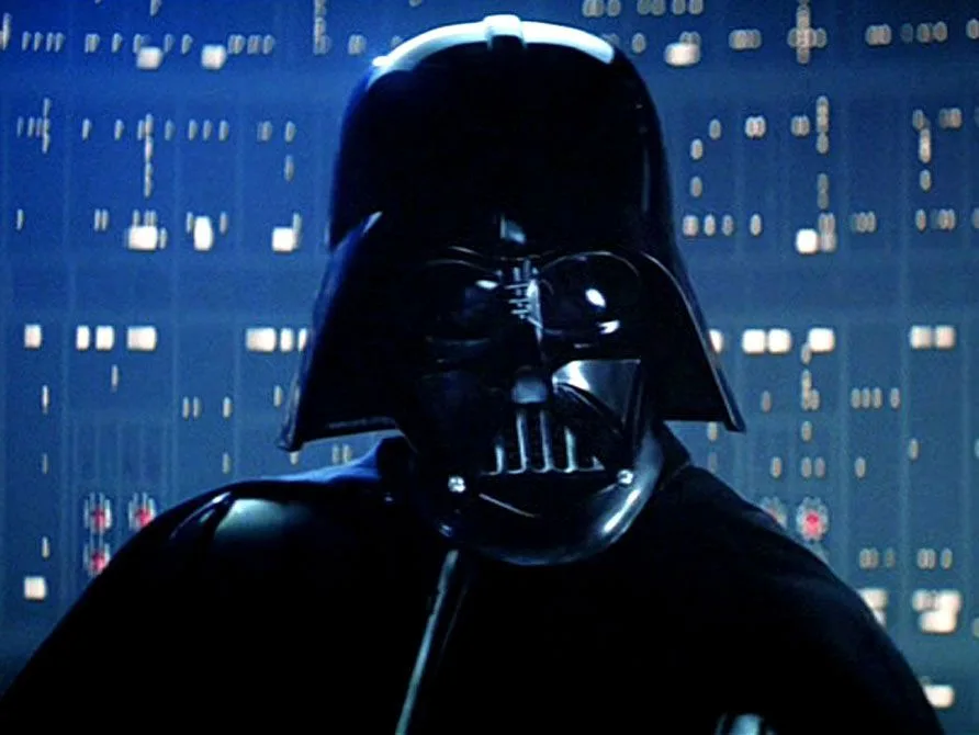 A close-up of Darth Vader in his iconic black helmet and suit, standing in front of a blue-lit control panel with glowing…