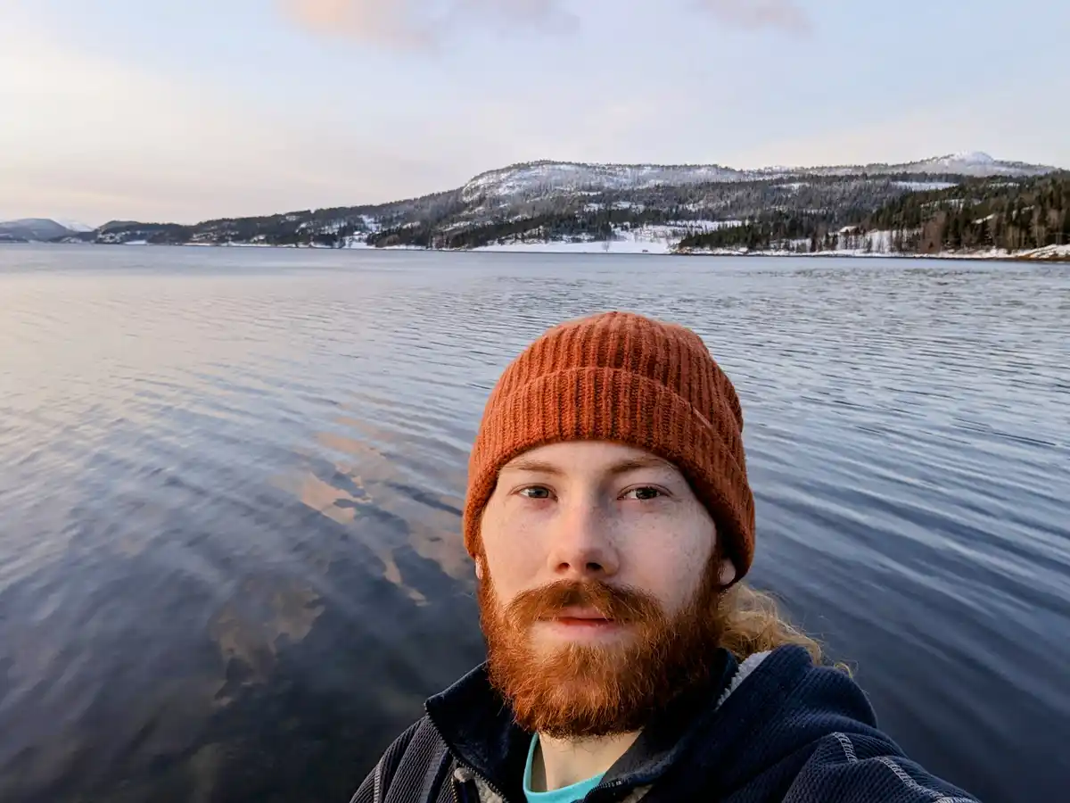 A man with a red beard and orange beanie takes a selfie by a calm, snow-dusted lake with mountains in the background. The…