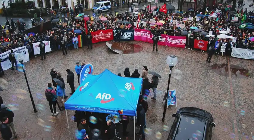 A large crowd of protesters holding signs and banners surrounds a blue AFD campaign tent in a wet cobblestone square, with…