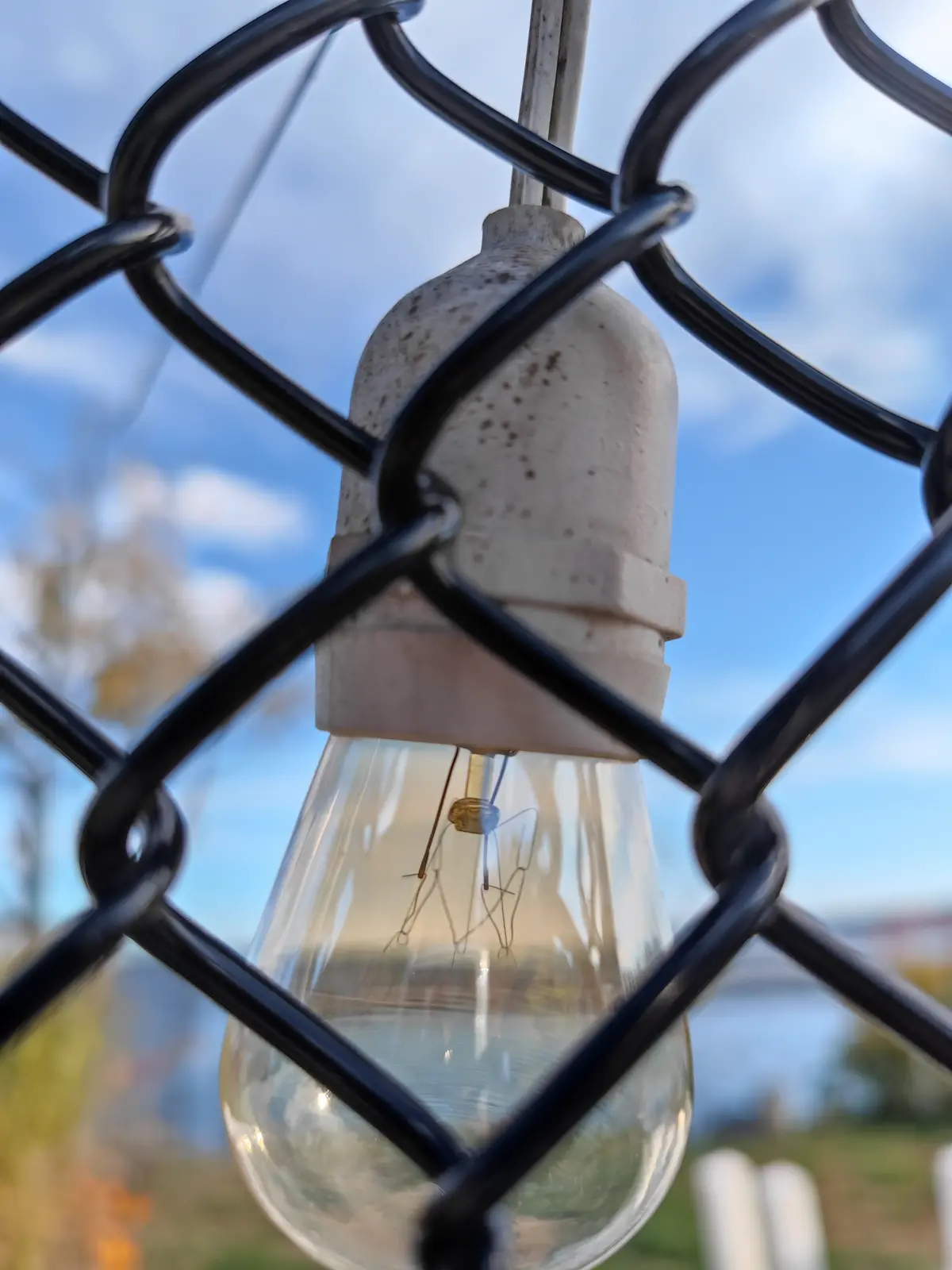 A close-up photo shows an unlit, clear glass incandescent light bulb hanging from a weathered, speckled ceramic socket…