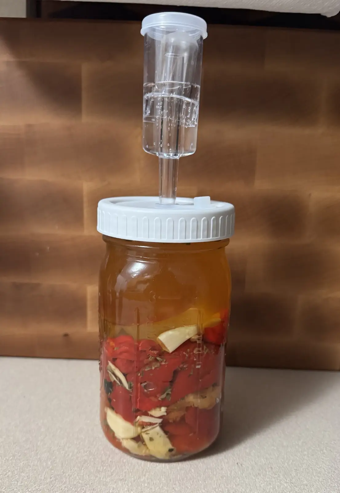 A glass mason jar filled with chopped red peppers, garlic cloves, and herbs submerged in liquid, topped with a white lid and…