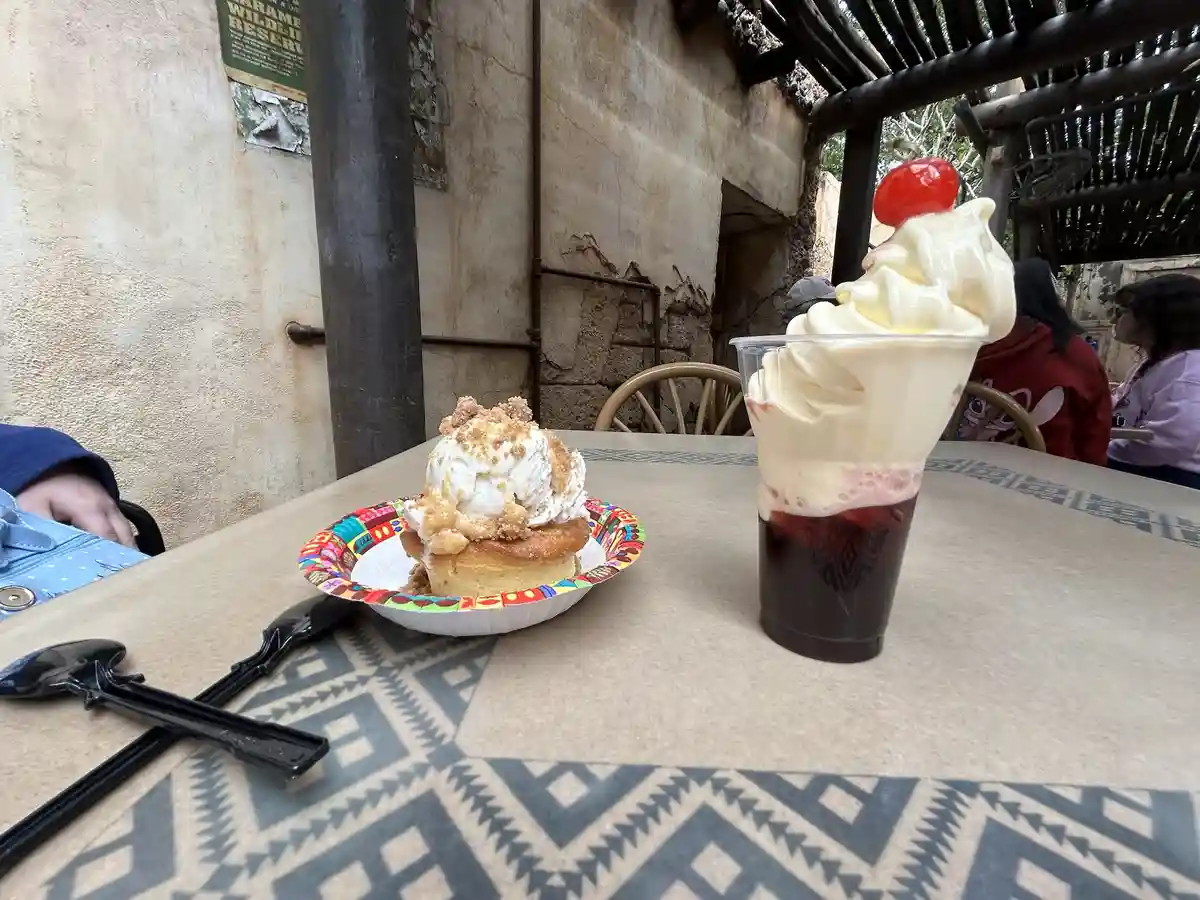 A table at an outdoor eatery features a dessert cup with whipped cream and a cherry on top next to a dessert on a colorful…