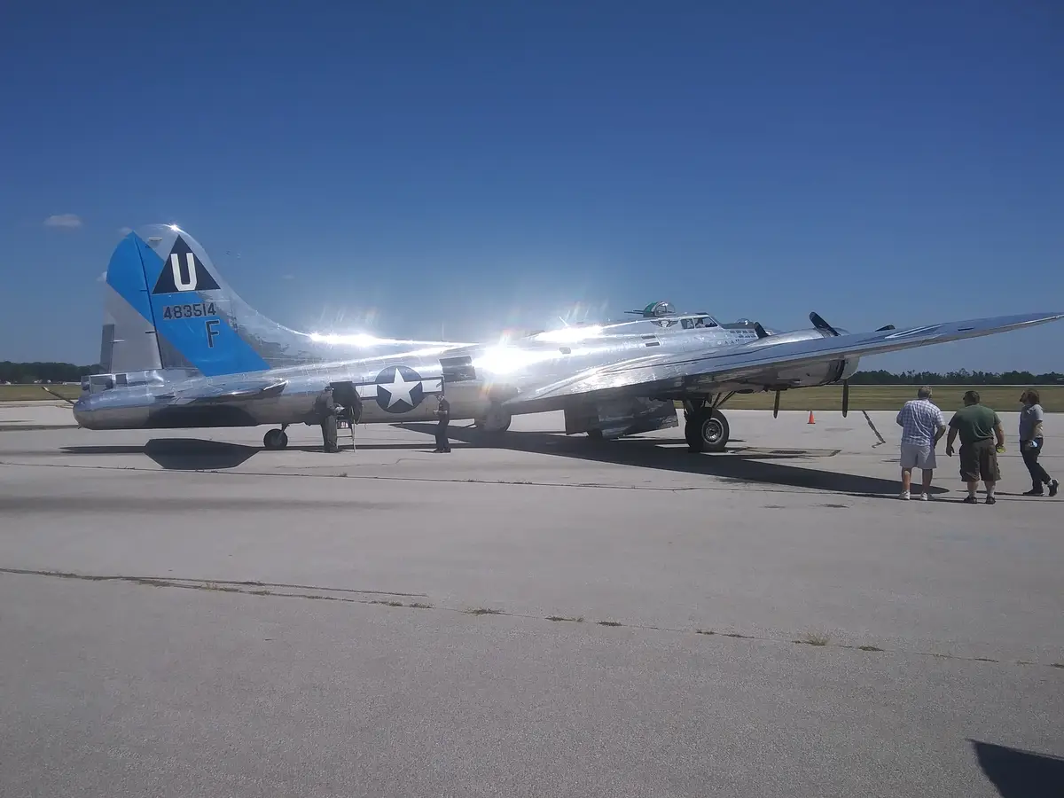 A shiny silver World War II-era bomber aircraft, likely a B-17 Flying Fortress, is parked on an airfield under a bright blue…