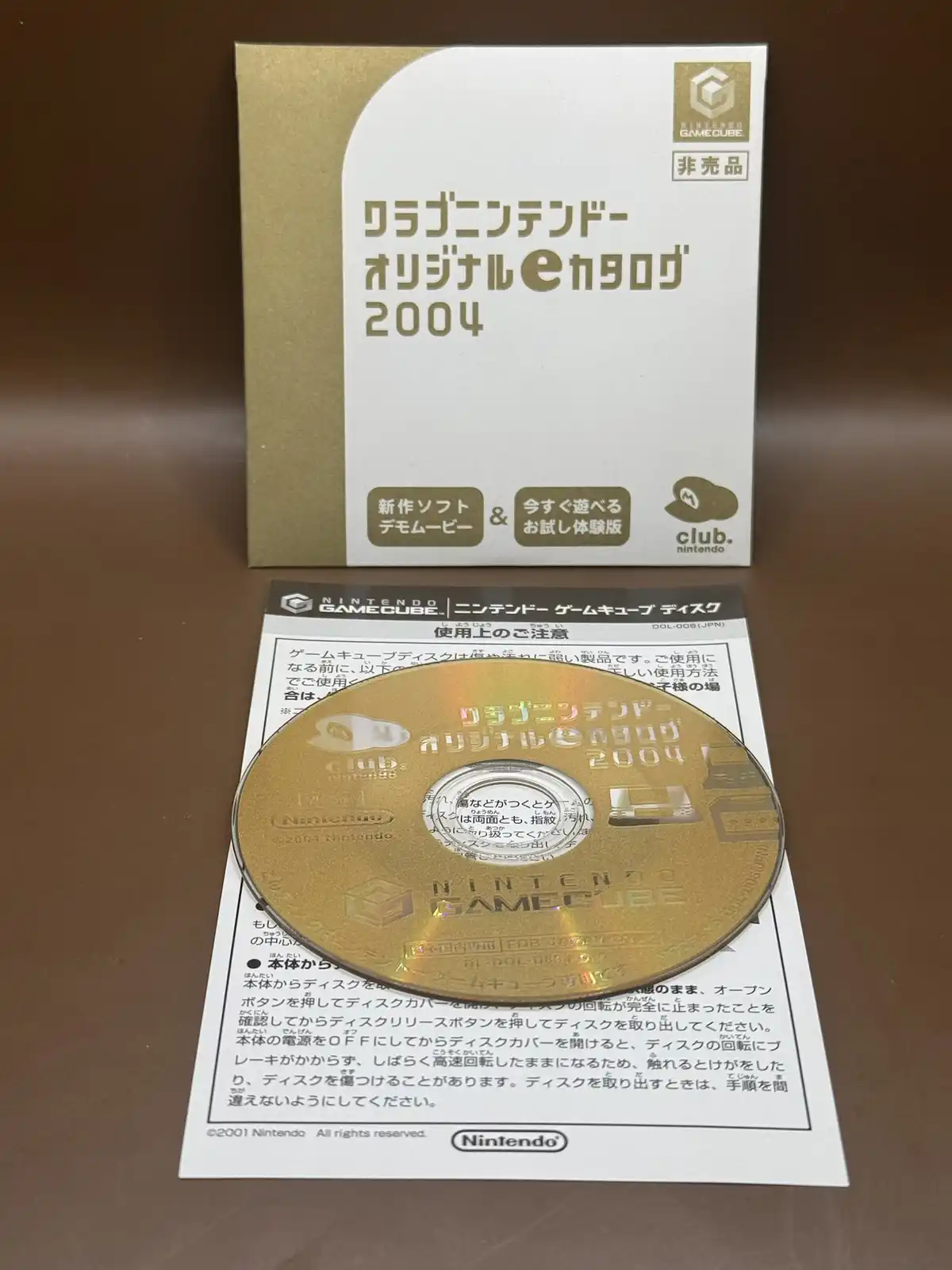 A gold Nintendo GameCube disc and its box for the 'Club Nintendo Original eCatalog 2004' are displayed on a brown surface…