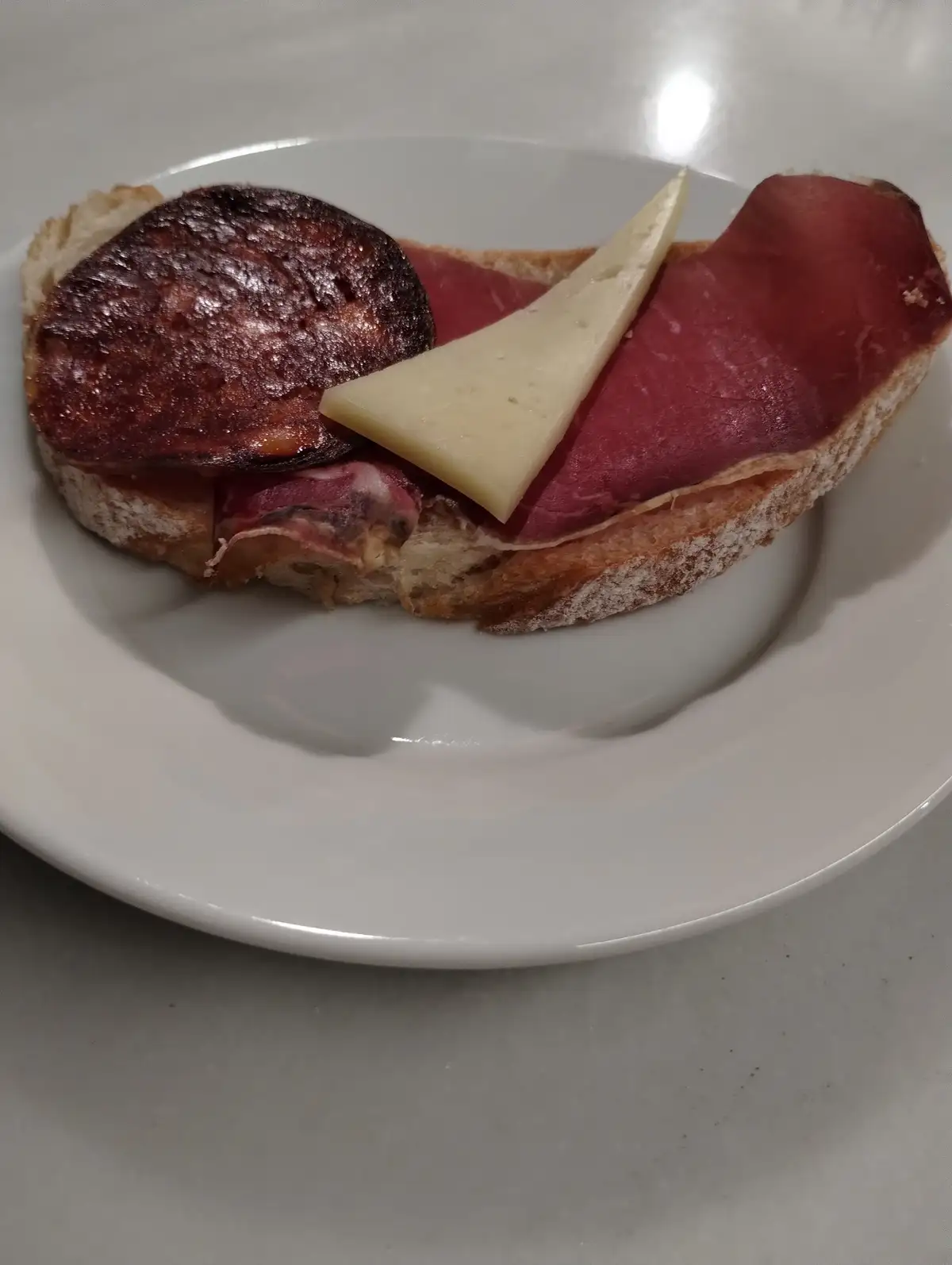a close-up photo of a sandwich on a white plate, featuring a slice of bread topped with a dark cured meat slice, a…