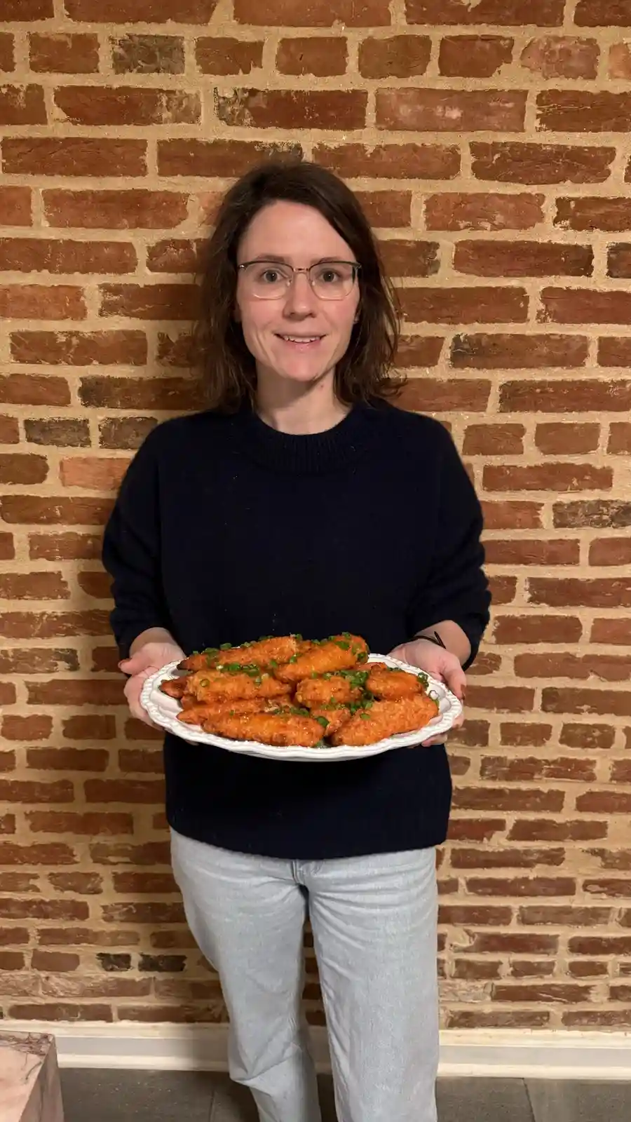 A woman with glasses and a dark sweater stands in front of a brick wall, holding a white plate piled high with golden-brown…