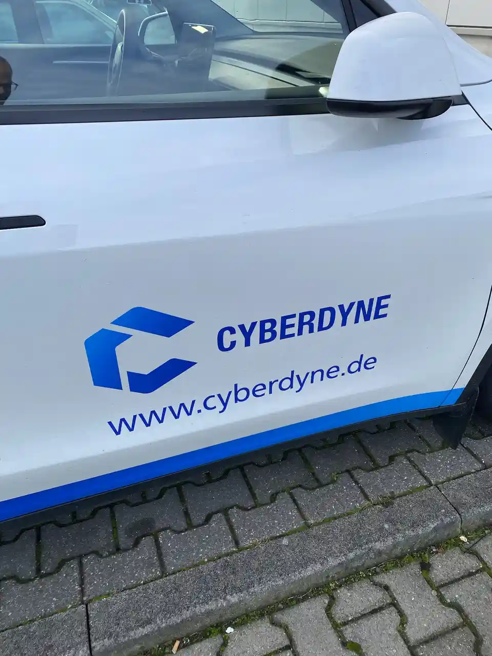 A close-up of a white car's side door showing a blue logo and text that reads 'CYBERDYNE' and 'www.cyberdyne.de'. The car is…