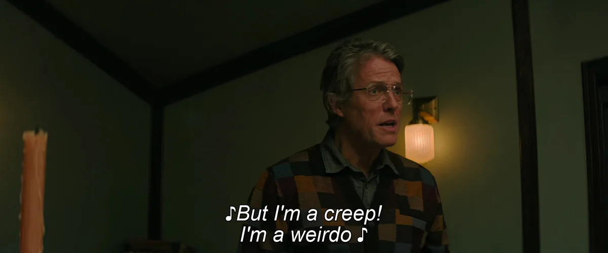A still from a movie shows a man with glasses and a plaid shirt looking surprised in a dimly lit room with a wall lamp…