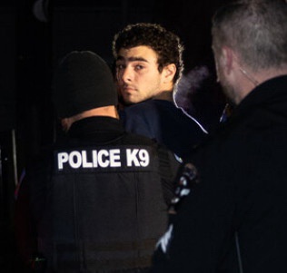 A man with dark curly hair and a surprised expression looks over the shoulder of a police officer wearing a vest labeled…
