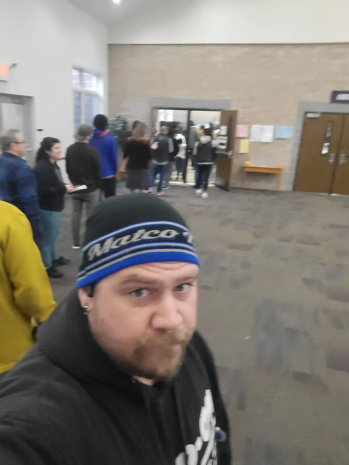 A man wearing a black beanie with 'Matco' written in white and blue stripes takes a selfie in a crowded indoor hallway. He…