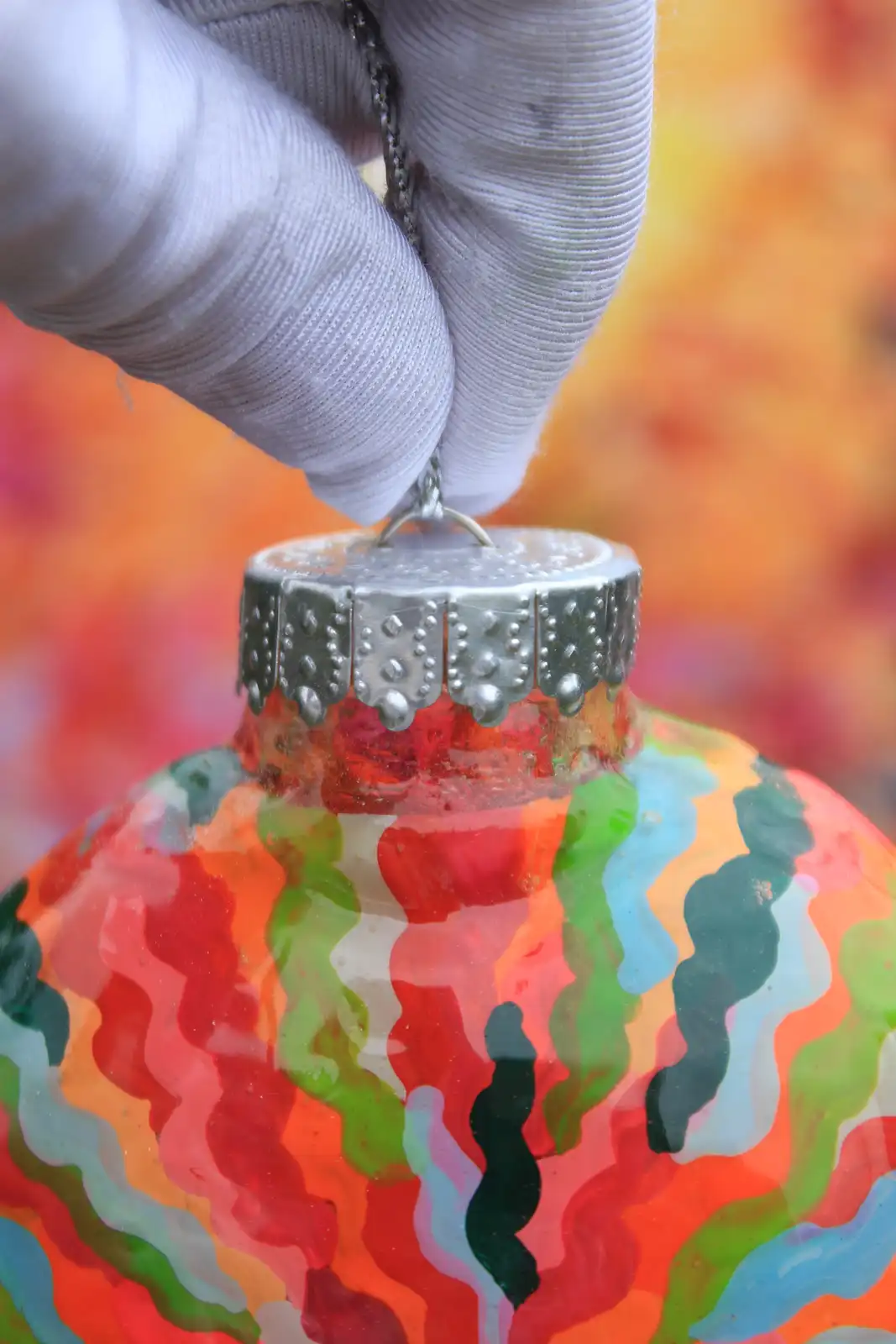 A close-up photo shows a hand in a white glove holding a colorful, hand-painted glass Christmas ornament by its metal cap…