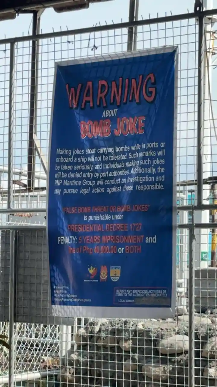 A blue warning banner is taped to a metal fence at what appears to be a port or construction site. The banner warns that…