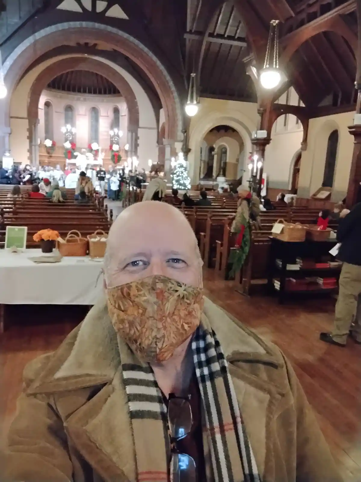 A bald man wearing a patterned face mask and a plaid scarf takes a selfie inside a decorated church with wooden pews, arched…