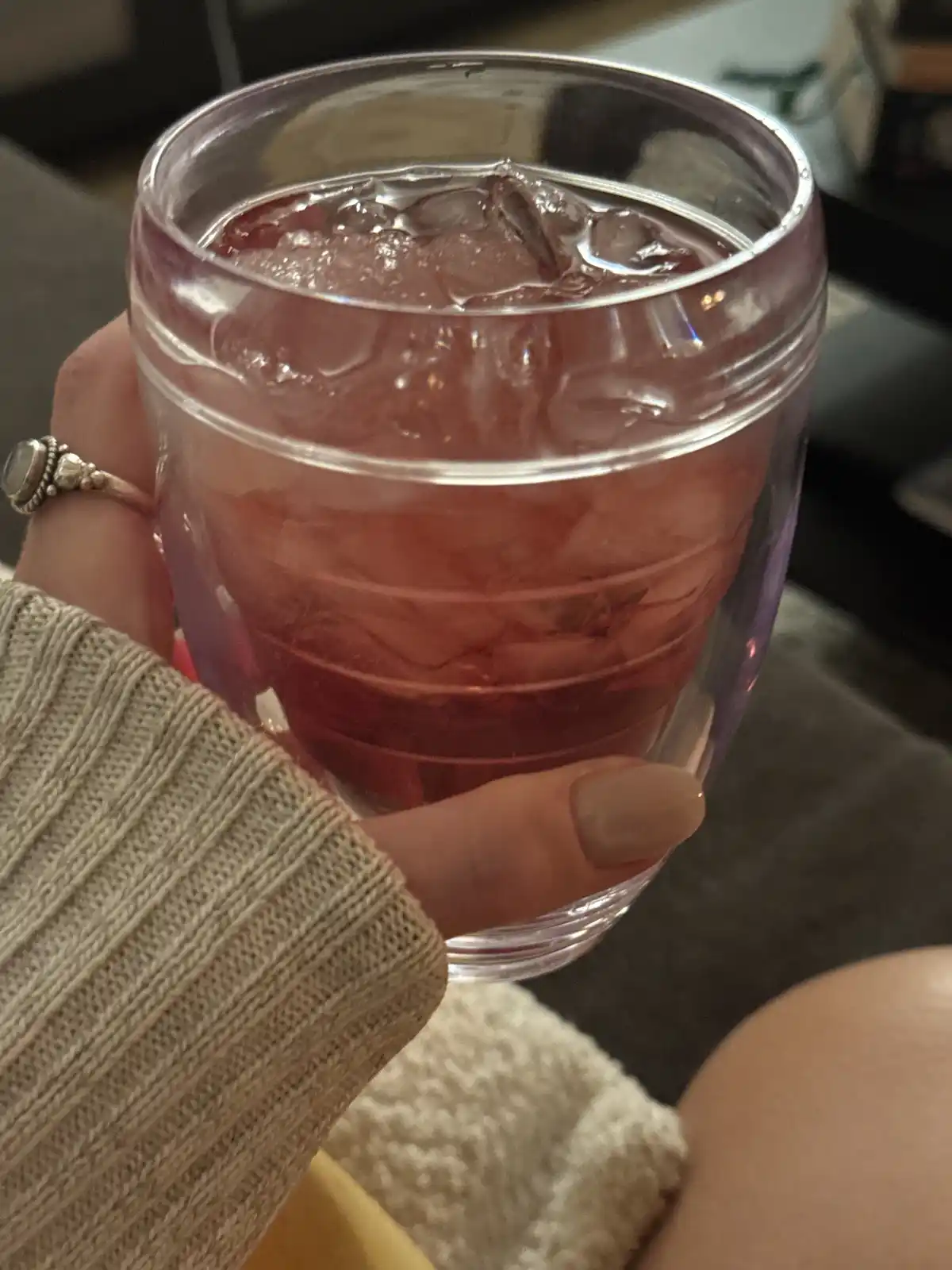 A close-up photo shows a hand holding a clear glass filled with a pinkish-red beverage and ice cubes. The hand, wearing a…