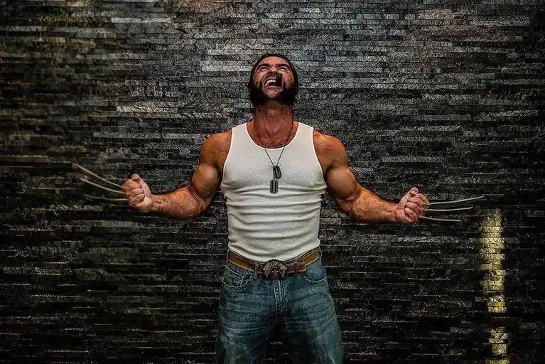 A muscular man with a beard, wearing a white tank top and jeans, stands against a dark stone wall with his arms…