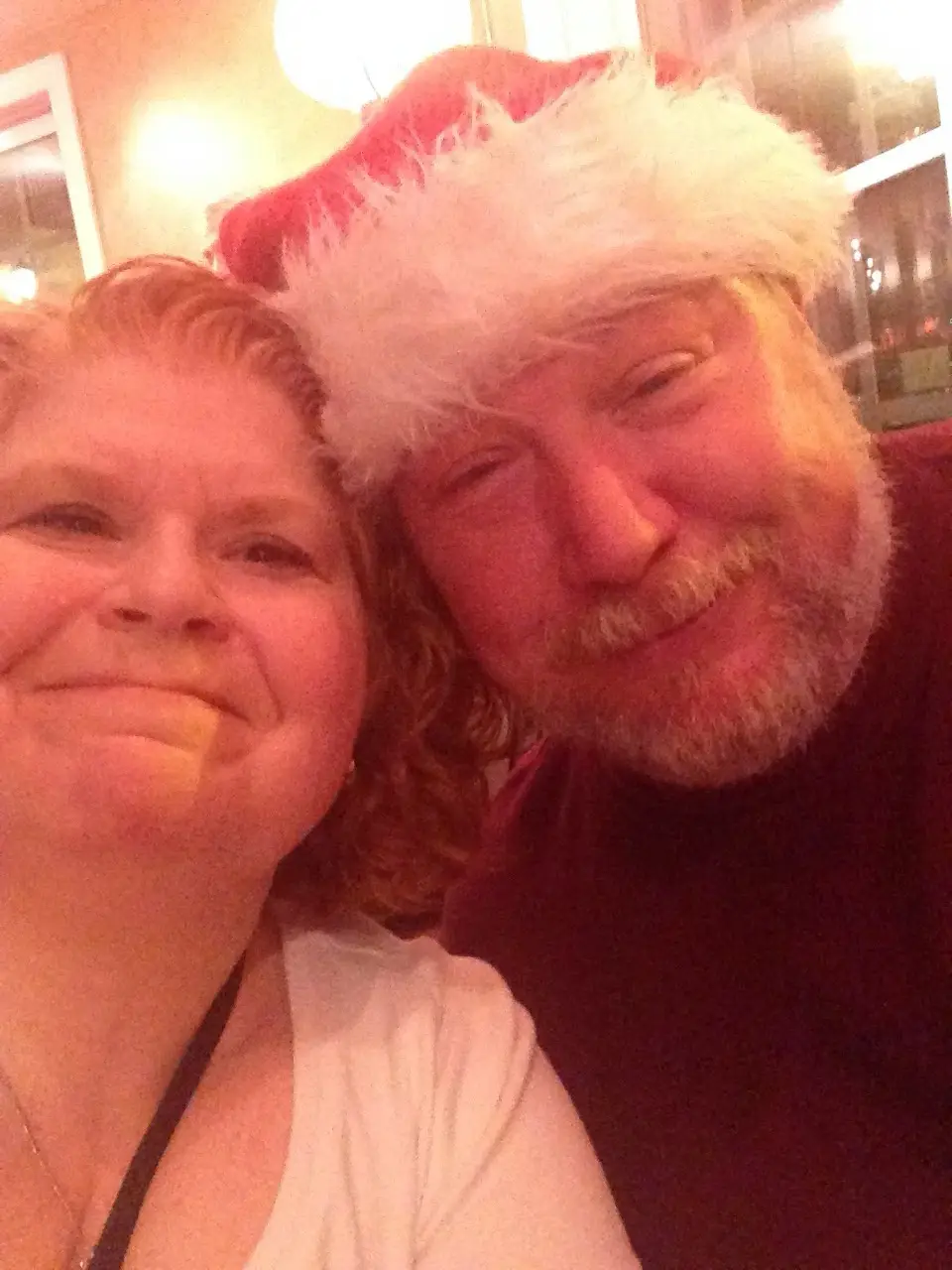 A selfie of a smiling man wearing a red and white Santa hat and a woman beside him, both looking at the camera. The man has…